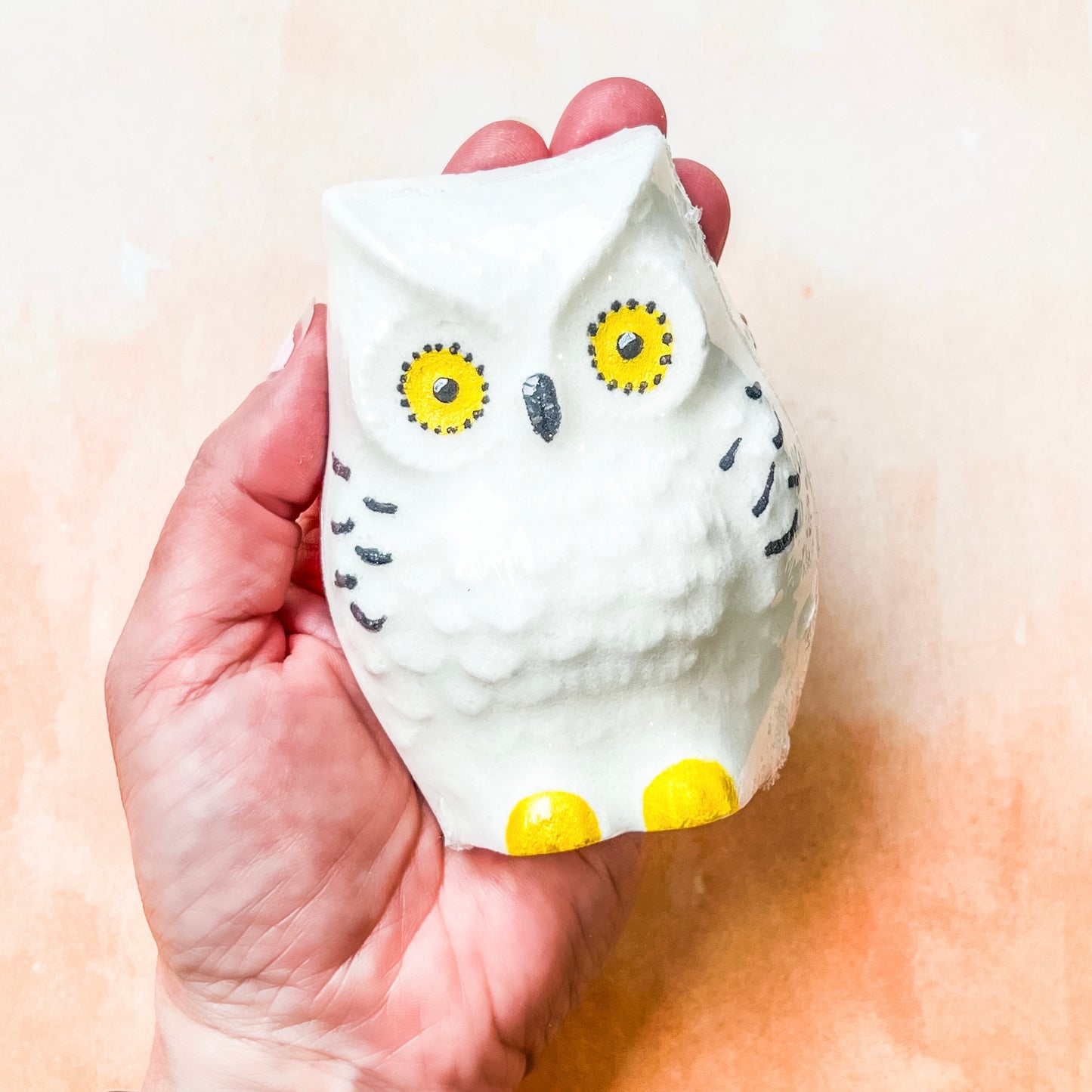 Hand holding a white owl-shaped bath bomb with yellow accents on a light background